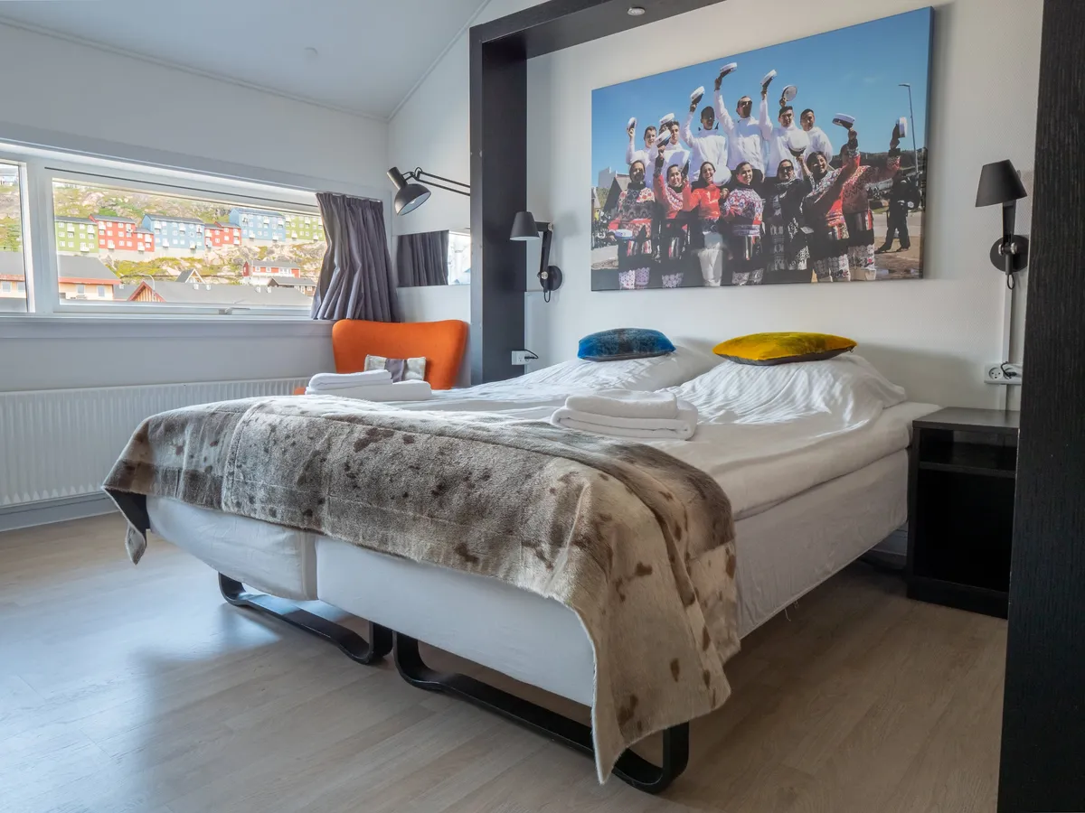 A double room: the beds under a fur throw, orange and yellow cushions, and a window over the coloured houses of Qaqortoq.