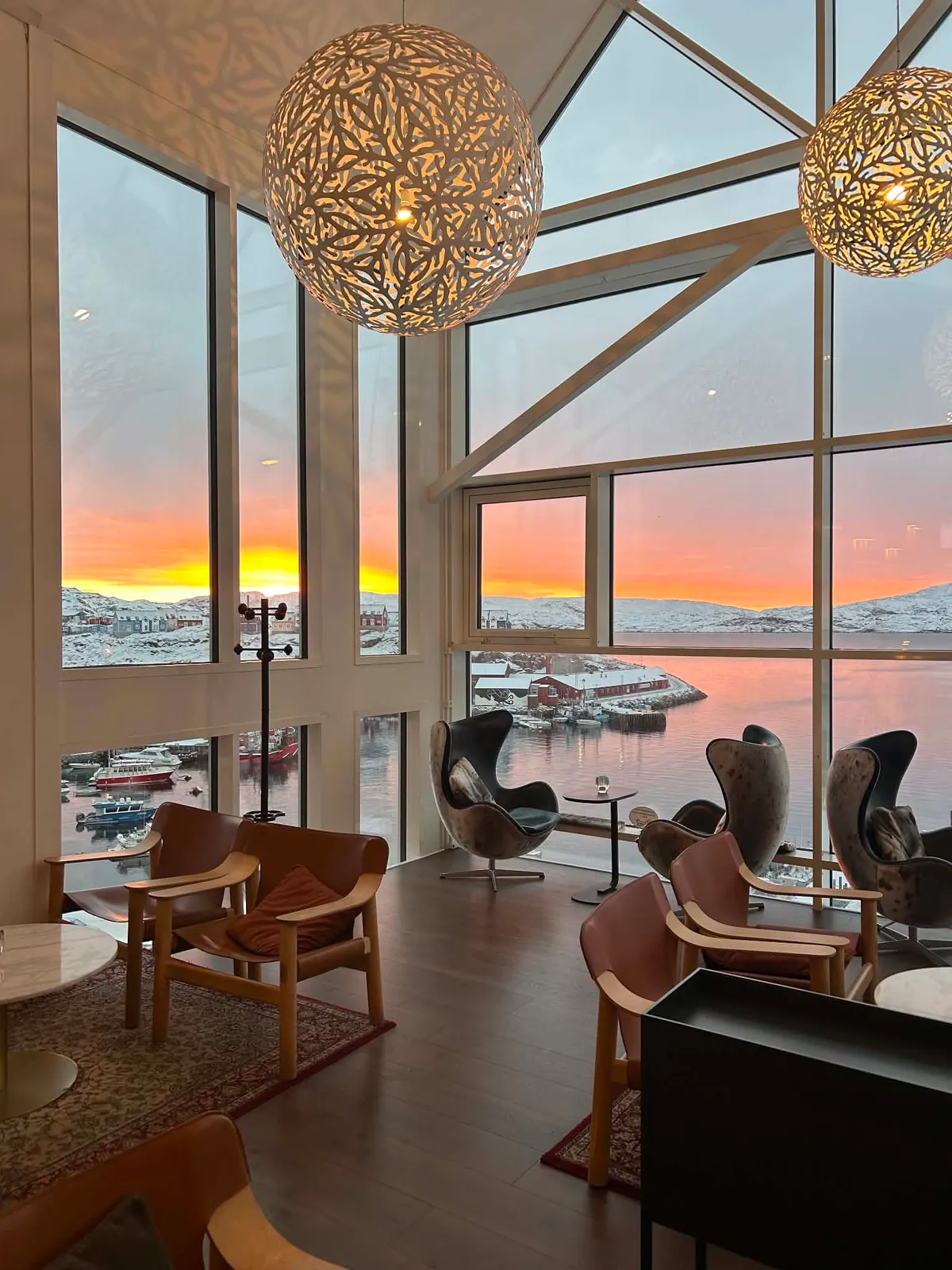The lounge at Hotel Qaqortoq at sunset: armchairs beneath tall windows looking out over the harbour and the snow-covered hills.